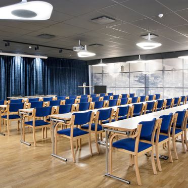 Konferensrum med vita långa bord, trästolar med blå dynor, blå gardiner, vita lampor i taket