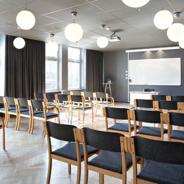 Konferensrum med ljus parkett, trästolar med grått tyg, grå gardiner i stora fönster, whiteboardtavla