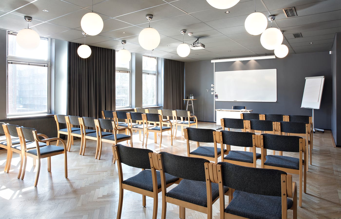 Konferensrum med ljus parkett, trästolar med grått tyg, grå gardiner i stora fönster, whiteboardtavla