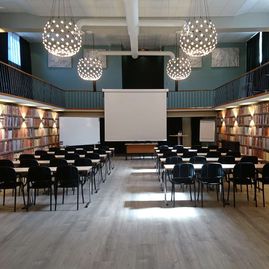 Rum med högt i tak, mörka stolar, väggar med böcker, stora lampor i taket