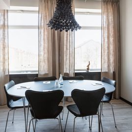 Mindre rum med ovalt bord, mörka stolar, lampa i taket, ljusa linnegardiner