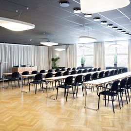Konferensrum med ljust parkettgolv, vita lampor i taket, långa vita bord, mörka tygstolar