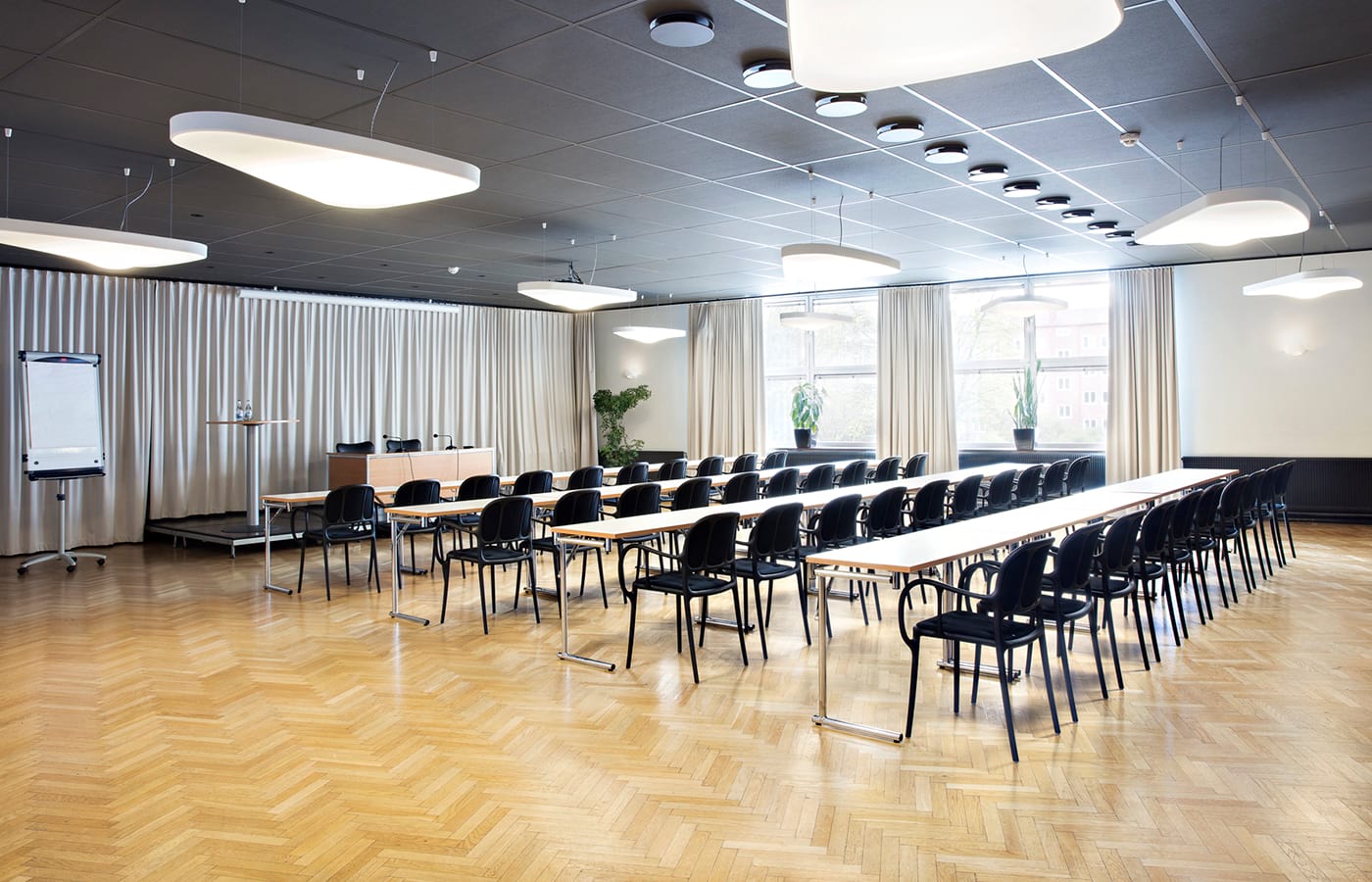 Konferensrum med ljus parkett, avlånga träbord med svarta stolar, vita lampor i taket, stora fönster med vita gardiner, stora gröna blommor.